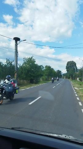 The media shows a view from inside a vehicle, looking out at a paved road with a solid white line on the left and dashed white lines on the right, indicating a two-lane road. The road curves gently to the left as it travels through a rural, tree-lined area. Two motorcycles are visible on the road, traveling in the same direction as the vehicle. The first motorcycle, closer to the camera, is black with orange accents and a rider wearing a helmet and dark clothing. The second motorcycle is white with a rider also wearing a helmet.

The setting is a rural landscape with lush green trees lining both sides of the road. In the background, hills or mountains are faintly visible. There are also signs of habitation, including fences and what appear to be houses or buildings on the right side of the road. A few power lines are visible strung across the sky.

The sky is a vibrant blue with fluffy white clouds, suggesting a pleasant day. The lighting indicates it is daytime, likely late morning or early afternoon. The overall mood is one of travel and exploration, possibly a scenic drive through the Romanian countryside. No text is clearly visible within the media file.