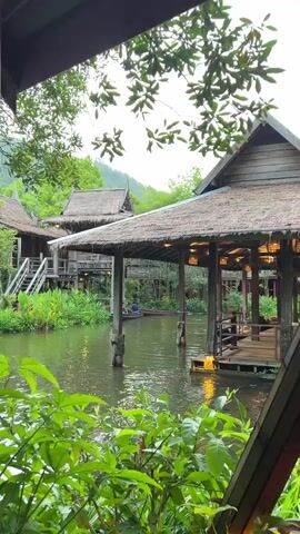 The video shows a serene canal with traditional wooden structures on either side, reminiscent of Southeast Asian architecture with their thatched roofs and elevated walkways. Lush green foliage frames the scene, with trees and plants in the foreground and background. The water is calm and reflects the surrounding structures and greenery. In the middle of the canal, a small wooden boat is being paddled by a person. The overall ambiance is peaceful and tranquil, suggesting a natural or resort-like setting. The lighting suggests it is either early morning or late afternoon, with soft, diffused light.