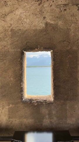 The video shows a view of a lake and mountains through a stone window. The window is rectangular and appears to be part of a stone wall. The lake is a light blue color, and the mountains are covered in snow. The sky is visible above the mountains. There is a dark colored table-like surface visible at the bottom of the window. The camera zooms in slightly to get a closer view.