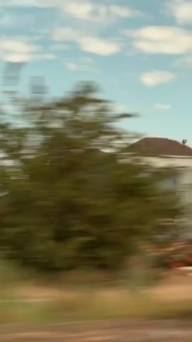 This video shows a blurry view of a landscape from a moving vehicle. The scene includes construction equipment, trees, a house, and a truck trailer. The sky is blue with some white clouds. The overall impression is of a rural or industrial area viewed at high speed, creating motion blur that obscures fine details but conveys a sense of movement and transit.