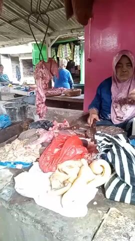 A woman wearing a pink hijab and a blue blouse sits at a market stall, chopping meat. The stall is covered with various cuts of raw meat, some in plastic bags. A large piece of meat hangs from a hook above the stall. In the background, another person is visible in a blue shirt. The market appears to be covered by a corrugated metal roof.