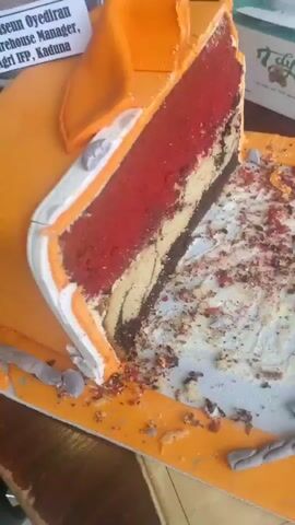 The media displays a close-up shot of a sliced cake, presented on a yellow cake board with some crumbs scattered around. The cake is elaborately decorated to resemble a shirt, with an orange fondant covering and white detailing on the collar and placket. A prominent slice has been removed, revealing multiple layers of cake in various colors: red, light yellow, and dark brown, with a marbled effect in the yellow and brown layers. The cake appears to be a centerpiece or part of a celebration.

Behind the cake, a photograph of a man wearing glasses and a white shirt is partially visible. Next to it, a printed card with text is propped up. The visible text on the card reads: "Oluwascun Oyediran, FG Warehouse Manager, Olam Agri Ire, Kaduna." This suggests the cake is a tribute or celebration for Oluwascun Oyediran, who holds the position of FG Warehouse Manager at Olam Agri Ire in Kaduna.

To the right of the cake, a box of tissues with a floral design is visible, hinting at a domestic or celebratory setting. The lighting suggests it could be daytime, possibly indoors. The presence of a sliced cake, a photograph, and a congratulatory card points towards a birthday, retirement, or farewell event. The overall mood conveyed is one of commemoration and perhaps a touch of sadness if it's a farewell. The location context of Gwagwada, Nigeria, places the scene within this specific Nigerian locale.