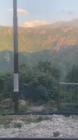 The video pans across a mountainous landscape during what appears to be either sunrise or sunset, casting a warm glow on the rocky peaks. In the foreground, there is evidence of human activity and possibly some neglect. A utility pole, painted with black and white bands, stands prominently in the center of the frame. To the left, a grey electrical or utility box is visible, mounted on a concrete base. The ground around it is littered with debris, including discarded plastic and metal objects, and patches of overgrown grass and weeds. Further to the left, a portion of a light-colored, angular building or structure is seen. Railroad tracks are visible at the bottom of the frame, suggesting the vantage point might be from a moving train. The overall impression is a blend of natural beauty and man-made elements, with a hint of disarray in the immediate surroundings.