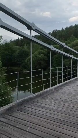 The video shows a perspective shot from a wooden bridge, moving forward. The bridge has metal railings and is suspended over water, which is visible below on the left. On the right, a dense forest with green trees lines the landscape, and the sky above is cloudy. The bridge leads towards an arched stone structure, a tower-like gate, with trees continuing behind it.