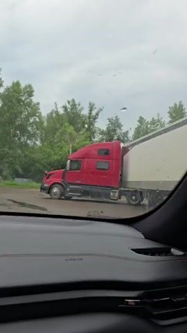 The video begins with a shot from inside a car looking out the windshield at a red semi-truck. The view then shifts to the interior of the car, showing a man driving. The camera pans to the dashboard, displaying a digital gauge and navigation screen. The video then returns to an exterior shot from the car's perspective, showing trees and a utility pole. Finally, the video shows a side-view mirror reflecting the car and the road behind it.