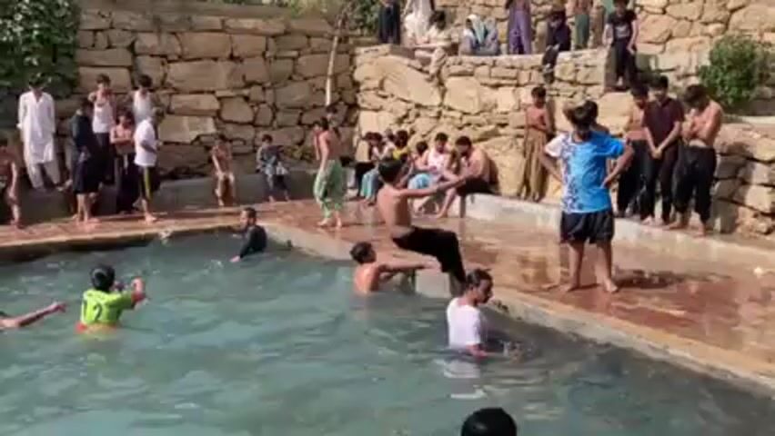 The video captures a group of young boys swimming and playing in a rectangular outdoor pool. The pool is surrounded by stone walls, with several people, mainly boys and men, sitting on the walls as spectators. Some of the boys are already in the water, while others jump in, creating splashes. The atmosphere appears relaxed and playful. The surrounding area includes a stone structure and what appears to be a natural landscape.
