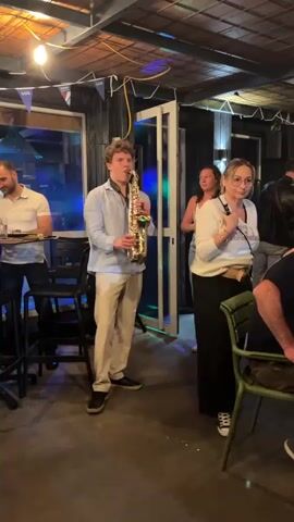 A young man with curly blonde hair is playing a saxophone. He is wearing a light blue collared shirt and cream-colored pants. He stands in front of a woman with blonde hair who is wearing a white sweater and dark pants. The woman appears to be singing or speaking into a microphone, and her mouth is open. In the background, another man in a white polo shirt stands near a bar. The scene appears to be an indoor venue, possibly a bar or restaurant, with string lights and flags hanging from the ceiling.