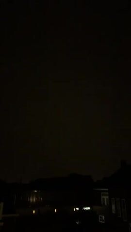 The video captures a dark night sky with a faint, intermittent glow. Below the sky, the silhouettes of buildings and trees are visible against the dim light. The overall scene suggests a low-light environment, possibly during a storm or with distant light pollution. There are no people or specific actions clearly discernible due to the darkness and low resolution.