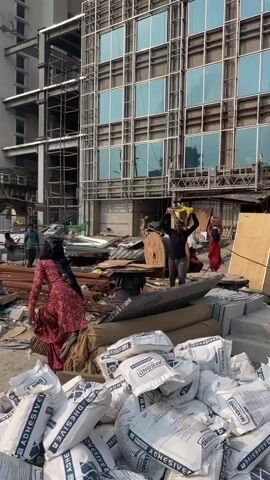 The video shows an active construction site in Noida, India, under bright daylight. In the foreground, numerous white bags of "MAXBOND ADHESIVE" are piled up. Behind them, various construction materials like rebar, large wooden spools, rolls of fabric or insulation, and concrete blocks are scattered across the uneven ground.

Several workers are visible. A man in a black shirt and grey pants is seen walking forward, carrying a yellow bundle or basket on his head with both hands raised to balance it. Another woman, wearing a red patterned dress, walks in front of him, also carrying a load on her head. In the background, other workers are seen amidst the debris and materials.

The backdrop features a partially constructed building with a prominent steel framework and several reflective blue-tinted glass windows already installed on its upper floors. Scaffolding is visible on the building's exterior. The overall scene depicts ongoing manual labor at a bustling urban development site.
