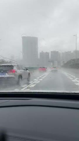 The video shows a view from inside a car driving on a highway during a heavy rainstorm. Water is streaming down the windshield, obscuring the view of the road and the surrounding environment. Several cars are visible on the road ahead, including a silver SUV in the left lane and red cars in the right lane. Tall buildings can be seen in the distance through the rain and fog. The road surface appears wet and reflective. The overall impression is of reduced visibility and difficult driving conditions due to the severe weather.