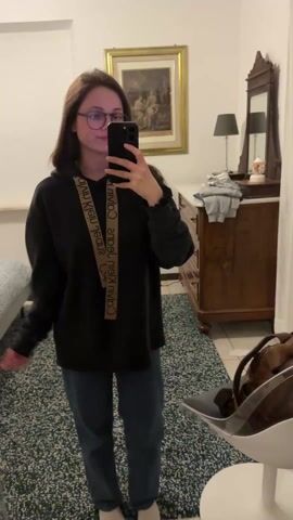A young woman is taking a selfie in a bedroom. She is wearing glasses, a black hoodie with a tan strap across it, blue jeans, and white socks. The room contains a bed, a dresser with a mirror, a painting on the wall, a lamp, a rug, and a clear plastic chair with a backpack on it. She is holding her phone in her right hand and looking at the screen, briefly puckering her lips.