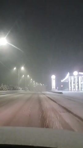 The video shows a car driving on a snow-covered road at night. It is snowing heavily, and the visibility is poor. Streetlights illuminate the road. A gas station is visible on the right side of the road. The car is moving forward.