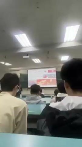 The video shows a classroom from the back, with students sitting at desks facing a projector screen. The projector displays a presentation, which includes images and text. The classroom has fluorescent lighting and a light green or blue-tinted desk surface. An instructor may be speaking in the front, but is not visible.