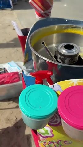 The video shows a cotton candy cart on a beach. The cart is decorated with cartoonish depictions of candy and the phrase "СЛАДКАЯ ВАТА" (Sweet Cotton). A steel bowl sits atop the cart, along with containers of colored sugar and a spray bottle. There is a clear acrylic shield around the bowl. The background shows a walkway and some beach furniture, and the sound of music can be heard.