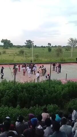 The video shows an outdoor court, possibly a tennis or volleyball court, with a chain-link fence surrounding it. There are several people on the court, most of whom are dressed in white or light-colored clothing. Some appear to be participating in an activity, while others are standing and observing. In the foreground, there is a dense area of green bushes and foliage, behind which a crowd of people is seated, facing the court. The background consists of a large, open, grassy field with scattered trees and utility poles under a cloudy sky. The lighting suggests it is late afternoon or early evening.