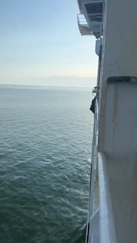 The media file shows a view from the side of a moving ferry. The ferry's white railing and rope are visible in the foreground. The sea is calm with small waves. The sky is clear and blue, suggesting a pleasant day. The sun appears to be high in the sky, indicating it is likely daytime. There are no visible people clearly in focus, although some figures can be seen on the deck of the ferry in the distance. The scene is peaceful and suggests a journey across the water. There is no visible text in the media file.