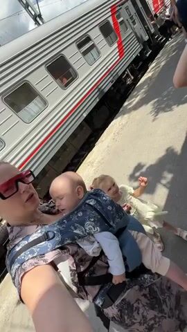 A woman is holding a baby in a carrier. A young child is running near her. A train is parked on tracks beside them. In the background, other people are standing. The scene appears to be a train station platform on a sunny day. The video then cuts to a close-up of a woman with a baby and then to a child looking at a store counter.