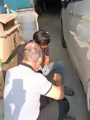 Two men are crouched down next to a silver car. The man in the foreground is wearing a grey polo shirt and black pants. The man behind him is wearing a black t-shirt and blue jeans. They appear to be working on the car's tire. The environment appears to be an outdoor area with a concrete floor and some boxes stacked in the background. Sunlight is casting shadows on the ground.