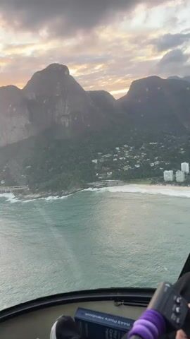 The video shows a first-person perspective from inside a helicopter flying over a coastal city at sunset. The camera pans from left to right, revealing a mountain range on the left, a coastline with a beach, a highway bridge, and buildings nestled among green trees. The sky is filled with clouds tinged with orange and pink hues from the setting sun. The helicopter's dashboard and a book with a purple water bottle are visible in the foreground.