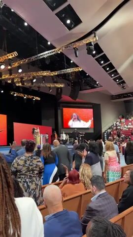 The video captures a lively scene inside a large indoor venue, likely a church or auditorium, filled with an audience seated on tiered benches. The focal point is a brightly lit stage with a vibrant red backdrop, featuring two large screens. On the stage, several individuals are present, some appear to be performers or speakers, with one person, dressed in a light-colored outfit, actively singing or speaking towards the audience. Another individual, dressed in dark clothing, is also on stage, seemingly interacting with the performer. The audience members are facing the stage, with many raising their hands, suggesting active participation in a service or performance. The venue itself is spacious, with a high ceiling equipped with elaborate lighting rigs and large speaker systems. The overall atmosphere is one of communal engagement and energetic worship or celebration.