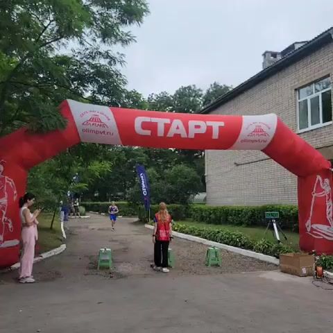 A male runner wearing a blue tank top and blue shorts runs towards an inflatable red archway with the word "START" written in white letters. There are two women standing near the archway, one wearing a pink shirt and light-colored pants, and the other wearing a red vest over a pink and white patterned shirt with black pants. A blue flag with white lettering is visible behind the runner. In the background, there is a brick building with windows, trees, and a green hedge. A tripod with a timer is placed on the right side of the path. There are also several green stools on either side of the path. The ground is paved with gravel and asphalt.