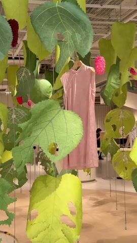 The video shows an indoor exhibition space. Large green paper leaves are suspended from the ceiling by wires, creating a canopy effect. Some of the leaves appear torn or have holes. In the center of the frame, a pink dress hangs from a hanger on a clothes rack. Several clusters of pink, berry-like objects are also visible, interspersed among the leaves. The background is slightly blurred, but appears to show display structures and possibly a few people moving in the distance. The lighting is bright, suggesting an exhibition or retail environment.