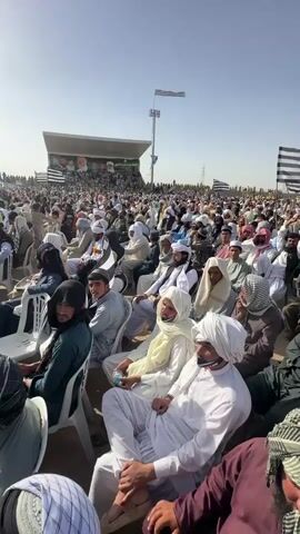 The video depicts a vast outdoor gathering of men, primarily dressed in traditional South Asian attire including shalwar kameez, turbans, and headscarves. The event takes place in a large, open field with dusty, arid ground under a clear, bright blue sky, indicating a sunny daytime setting.

Thousands of attendees are visible, with many seated on white plastic chairs in the foreground and stretching far into the background. In the distance, a large stage with a banner is set up, suggesting a public event like a political rally or religious congregation. Numerous black and white striped flags, commonly associated with the Jamiat Ulema-e-Islam (F) political party in Pakistan, are prominently displayed throughout the crowd and on tall poles. Power lines and utility poles are discernible in the far background.

The men appear to be attentively listening or observing the proceedings from the stage. Several individuals are holding plastic water bottles. The overall scene portrays a large, organized gathering in Pishin, Pakistan, characterized by a sense of order and engagement from the crowd.