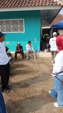 The video opens with a medium shot of two men sitting on the ground in front of a turquoise building. The man on the left wears a brown patterned cap and a brown shirt, while the man on the right wears a white shirt and light-colored pants. A motorcycle is parked to the left of the frame.

The camera then pans to the right, revealing a young woman in the foreground wearing a red hijab, a white long-sleeved shirt with red lettering on the back, and blue flared jeans. She has a brown shoulder bag. She is facing away from the camera and appears to be observing an event taking place under a blue tent in the background.

Several other individuals are visible under the tent. One man in a white shirt and a red and white headscarf is holding a microphone and speaking. Another person in a pink dress and a grey vest is standing near him. Other people are milling around in the background, some in white shirts. The environment appears to be an outdoor gathering, possibly a community event or a political rally, with a dirt ground and several buildings visible. The overall lighting suggests it is daytime.