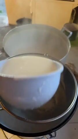 This is a video capturing a domestic scene in Starokhalilovo, Russia. The primary subject is the process of straining a white, liquid substance, likely a dairy product like cheese or yogurt, through a fine-mesh sieve. The sieve is placed over a large white enamel bucket, which is resting on a table or countertop.

In the foreground, a white plastic colander is held by an unseen hand, pouring the liquid into the sieve. Drops of the liquid are visible clinging to the colander. Behind the sieve, a large pot or bowl with a steaming, pale liquid is visible, suggesting that the substance being strained was recently heated.

The setting appears to be a kitchen or food preparation area. The background is somewhat blurred but includes what looks like a gas stove and possibly some boxes or storage containers. The lighting suggests it is daytime, possibly late afternoon or early evening, given the warm, soft light. There are no clear indications of weather or strong emotions, the focus being on the task at hand.

The activity is a step in food preparation, focusing on separating curds from whey or refining a liquid mixture. The scene conveys a sense of domesticity and hands-on cooking. No visible text is present in the video.