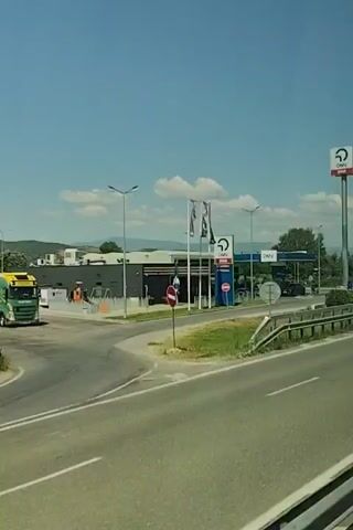 The video shows a view from a moving vehicle looking towards a gas station with OMV branding. A tall OMV sign is visible on the right. The gas station has multiple pumps under a blue canopy. Several cars are parked at the pumps. The gas station is located next to a highway, which is also visible in the foreground. The weather appears to be clear with a blue sky and some clouds. Trees and hills are visible in the background.