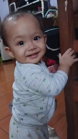A baby in a white onesie with a light green pattern is looking at the camera, smiling. The baby is in a room with a tiled floor and a wooden chair. The camera is held by someone who is out of focus in the foreground.