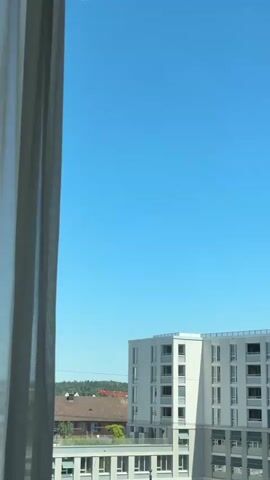 A white airplane with red accents flies across a clear blue sky, viewed from a window. The airplane is positioned in the upper part of the frame. Below the airplane and the sky, a modern white building with multiple windows is visible. To the left of the building, there are some rooftops with red tiles and trees. The left edge of the frame is partially obscured by a sheer, light-colored curtain. The camera is stationary and the airplane moves from left to right across the sky and then out of frame.