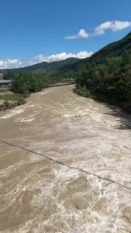 This video captures a wide, muddy river flowing through a valley under a bright, partly cloudy blue sky. The river is churning with rapids and white water, suggesting a strong current. The banks of the river are lined with green trees and vegetation. In the background, rolling hills covered in dense green forests rise up, and a few small buildings are visible on the left bank, suggesting a rural or semi-rural setting near a settlement. A bridge with a reddish-brown railing spans the river in the mid-ground. A thin, dark cable runs across the foreground of the image, likely part of the bridge structure or another overhead utility line. The overall impression is a scenic, natural landscape experiencing a powerful flow of water, likely due to recent rainfall or snowmelt. The time of day appears to be daytime, with bright sunlight and distinct shadows. The weather is fair and sunny with scattered white clouds. There is no visible text in the media.