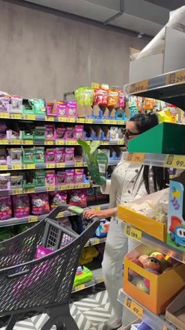 A woman with long dark hair pulled into a high ponytail is in a grocery store, pushing a shopping cart. She is wearing a white jacket, light-wash jeans, and dark sunglasses. She is holding a green potted plant in her right hand, examining it. The shopping cart is dark gray and appears empty.

The shelves in the background are stocked with various products, many of which are bagged or boxed pet food, indicated by the images of cats and dogs on some packaging. The aisle has a patterned floor with black and white zig-zag lines. An exit sign with Russian text ("ВЫХОД") is visible on the wall above a doorway. The lighting suggests it is daytime. The overall impression is of a routine shopping trip.