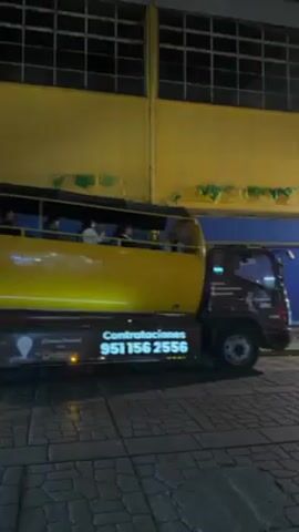 Here's a matter-of-fact description of the video:

The video shows a yellow bus, possibly a party bus, driving slowly on a cobblestone street at night. People are visible inside the bus, some appear to be looking out the open side windows. The bus has advertising, including a phone number.  A large yellow building with a grid of windows is in the background. The lighting suggests it's nighttime with artificial light illuminating the scene. There is audible cheering in the background.