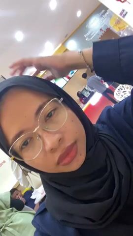 The video is a first-person perspective of a young woman wearing a black hijab and glasses. She is indoors, in what appears to be a cafe or restaurant, indicated by the tables and other patrons visible in the background. The lighting is bright, suggesting overhead fluorescent lights. The woman is looking directly at the camera and occasionally touches her forehead and adjusts her glasses. In the background, other people are seated at tables, some looking at their phones. The overall atmosphere seems casual and relaxed.