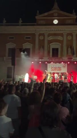 A nighttime outdoor concert is taking place in front of a large, ornate building. A band named "Tarraco Surfers" is performing on a brightly lit stage, with red lights and smoke effects. The band members are varied in their attire and appear to be dancing and interacting with the crowd. A large crowd of people are gathered in front of the stage, many of whom are holding up their hands and appear to be enjoying the performance. The building behind the stage has a clock tower and classical architectural elements. The overall atmosphere is festive and energetic.
