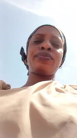 A woman is facing directly at the camera. She is wearing a patterned head wrap and a beige colored top. She has a serious expression. The background is light blue, suggesting it is outdoors with a clear sky.