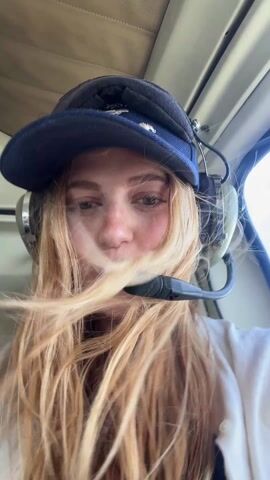 A young woman with long blonde hair and freckles is wearing a blue baseball cap and a headset with a microphone. She is inside an aircraft, possibly a helicopter, and looks into the camera with a smile. The camera moves around to focus on her face from different angles. The aircraft's interior is visible in the background.