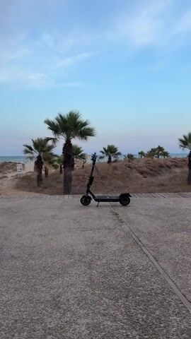 The video begins with a view of an electric scooter parked on a concrete path. Palm trees and dry vegetation are visible in the background, with the ocean and a clear blue sky beyond. A person's hand enters the frame from the left, holding a cigarette, and then moves out of frame. The camera then zooms in slightly on the scooter, which remains stationary. The scene appears to be a coastal area, possibly during the late afternoon or early evening, given the soft lighting.