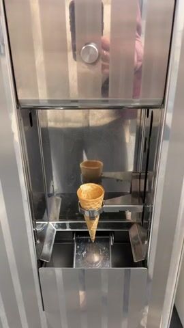 The video shows an ice cream machine dispensing soft serve ice cream into a cone. The machine is made of stainless steel with vertical silver stripes. An empty waffle cone is initially held in place by a silver clamp within the dispensing area. A person's reflection is visible in the machine's surface, showing them holding a phone and wearing a light pink sweater over a white shirt. The person is positioned in front of the machine, presumably operating it. As the video progresses, the ice cream machine dispenses a swirl of white soft serve ice cream into the cone. The ice cream slowly fills the cone from the top down. The video ends with a fully dispensed ice cream cone.