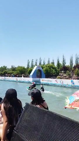 The video shows a water-based event from the perspective of someone standing near a crowd of people, overlooking the event. There is a lagoon with an obstacle course set up, featuring a large blue and white archway in the background that includes a Red Bull logo. The lagoon has individuals participating in the event on watercrafts. There are crowds of spectators along the sides of the lagoon watching the watercrafts. The sky is clear and blue. The camera pans back to show the crowd of people, mainly in the back, wearing casual clothing.