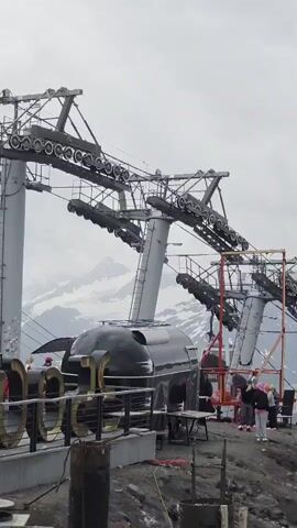 This video shows a ski lift station with several gondolas. In the foreground, large gold-colored letters that appear to spell "ELBRUS" are visible. A black, bullet-shaped structure with glass windows, possibly a station building or a specialized gondola, is also prominent. The ski lift towers and cables are complex and industrial, with visible wheels and mechanisms. In the background, snow-capped mountains rise against a cloudy sky, indicating a mountainous, possibly cold environment. Several people are present near the black structure and the ski lift, some appearing to be workers or waiting passengers. One gondola is in motion, descending from the upper part of the ski lift. The overall scene suggests an operational or recently completed ski resort infrastructure in a high-altitude location.