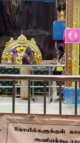 Inside what appears to be a Hindu temple or shrine, a man and a woman are engaged in a religious interaction. The setting features a richly decorated golden altar-like structure in the background, adorned with flowers and representations of deities. The man, likely a priest or temple officiant, is bare-chested and wears a traditional cream-colored dhoti. He stands behind a low metal table, facing the woman.

The woman, dressed in a floral long-sleeved top and dark pants, stands opposite him, holding a yellow bag. The man extends his hands, seemingly offering a blessing or an item (like prasadam) to the woman, who receives it with her hands clasped together in a respectful gesture. The interaction appears calm and reverent.

The background shows natural rock formations, suggesting a cave temple environment. To the right, there's a blue box and a pink sign with a white yin-yang symbol. Metal railings separate the foreground from the main area of interaction.

At the very bottom of the frame, a banner with Tamil text is visible. The text reads "கோயில்களுக்குள் கால் அணியக்கூடாது", which translates to "Footwear should not be worn inside temples". This sign further emphasizes the sacred nature of the location, consistent with the context of a Hindu temple in Malaysia, possibly within the Selayang Municipal Council area.