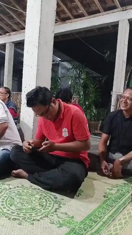 At 00:00, the video begins by showing a man in a red shirt and black pants sitting cross-legged on a green mat, looking down at a smartphone in his hands. He is wearing glasses. To his left, another man in a white and blue striped shirt and jeans is also sitting cross-legged, looking away from the camera. Behind them, other people are visible in the background, seated and blurred. The environment appears to be an outdoor or semi-outdoor covered area with a thatched roof supported by concrete pillars. At 00:03, a person's arm enters the frame from the top, holding a tray with food containers. At 00:04, the video cuts to a close-up of a person's legs and lower torso wearing a dark purple top and black pants.