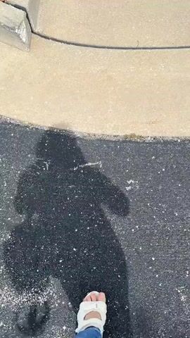 A first-person perspective video shows a person walking on a sidewalk. The person is wearing white sandals and blue jeans. Their shadow, which appears to be of a person with long hair holding a bag, is visible on the concrete. The sidewalk is next to a black surface, possibly asphalt, and there is a curb on the left side of the frame. The video is filmed during the daytime, as indicated by the shadows and bright lighting.