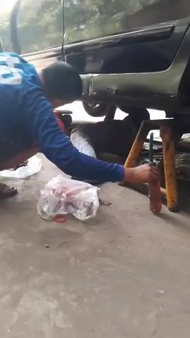 A person is working on the underside of a black car that is propped up on yellow jack stands. The person is wearing a blue long-sleeved shirt, dark patterned shorts, and flip-flops. They are crouched down and holding an orange bottle, possibly spraying something onto the car's undercarriage. Another person's feet, wearing flip-flops, are visible on the left side of the frame. A plastic bag containing what appears to be red items and a red rubber band lies on the ground in front of the person working on the car. The ground is concrete, and there are some tools scattered around. Trees and foliage are visible in the upper part of the frame, suggesting an outdoor or semi-outdoor setting.