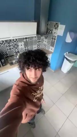 A person with curly dark hair is standing in a kitchen and filming a video. The camera rotates around, focusing at times on the person's face and hair. The person is wearing a brown hoodie with gold lettering and jeans. The kitchen has white cabinets, a patterned backsplash, and a white tiled floor. The walls are painted in shades of blue. A microwave and other kitchen appliances are visible.