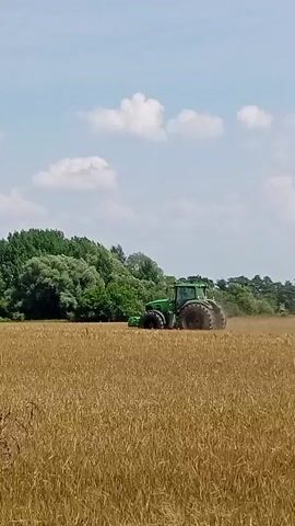 A green tractor is shown in a field of wheat under a blue sky with white clouds. The tractor is moving from left to right across the frame, kicking up dust as it goes. There are trees in the background.