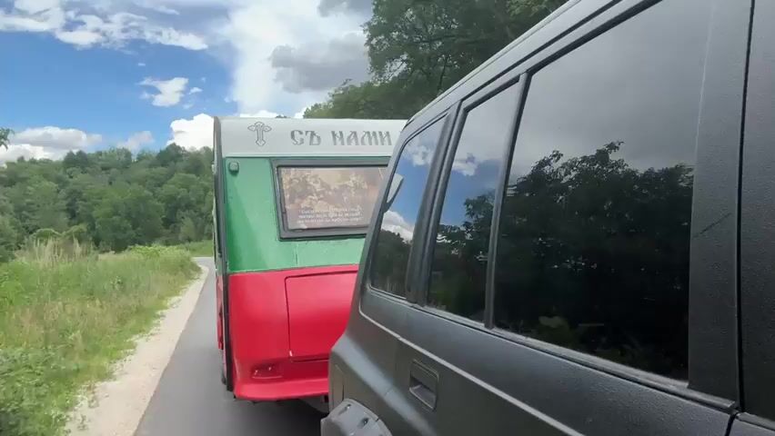 The video shows a close-up perspective from inside a dark-colored SUV, looking back at a trailer being towed. The trailer is brightly colored with red on the bottom and green on the top. It has a window with a religious image and text visible above it. The text appears to be in Cyrillic script and reads "СЪ НАМИ" which translates to "With Us". The trailer is being towed down a paved road, which is flanked by lush green vegetation and trees on either side. The road curves slightly to the left, and a white marker post is visible on the left side of the road. In the background, rolling hills covered in dense forest stretch towards a bright blue sky with scattered white clouds. The overall impression is of a journey through a natural, possibly rural, landscape. The time of day appears to be daytime, judging by the bright sunlight and the shadows. The weather seems fair, with a mix of sun and clouds. The scene evokes a sense of movement and travel through a scenic environment. The specific location is Biaga, Bulgaria.